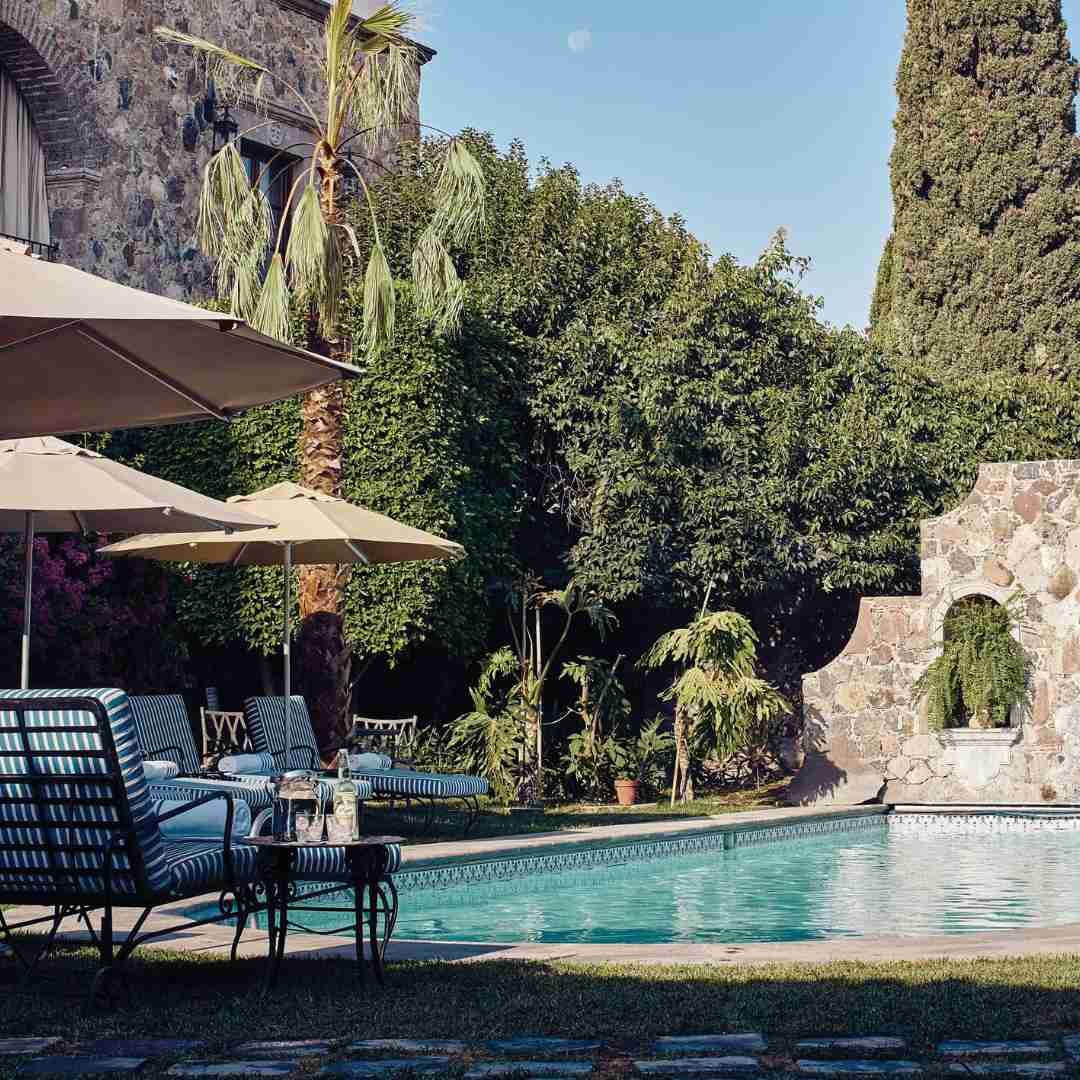 A tranquil photograph showcasing the pool at Belmond Casa de Sierra Nevada in San Miguel de Allende, Mexico. The crystal-clear water reflects the blue sky, surrounded by lush greenery and elegant lounging spaces.