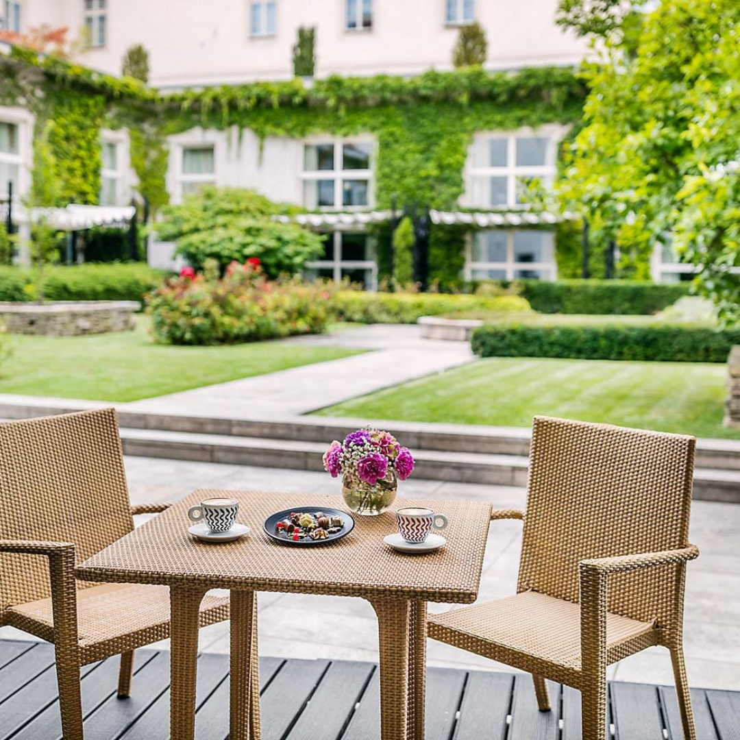 A tranquil scene in the gardens of Mandarin Oriental Prague, Czech Republic. A breakfast table is set in the foreground, surrounded by lush greenery and the hotel's verdant walls. The serene garden provides a picturesque backdrop, inviting guests to savor a peaceful and luxurious dining experience in the heart of Prague.