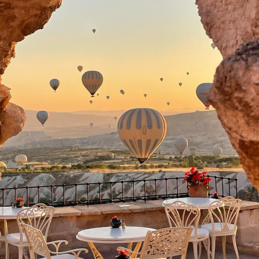 A captivating scene at the Museum Hotel in Cappadocia, Turkey. The restaurant boasts tables in the foreground, offering a front-row seat to the breathtaking Cappadocian landscape. In the background, hot air balloons paint the sky, creating a picturesque setting for guests to enjoy a dining experience surrounded by the magic of Cappadocia.