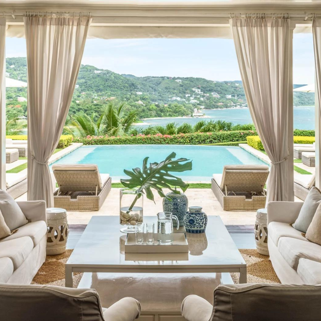 A scenic view of Round Hill Hotel in Montego Bay, Jamaica. The luxurious lounge and pool area beckon, surrounded by tropical beauty. In the background, the mesmerizing Caribbean Sea extends to the horizon, adorned with an island vista. A captivating blend of elegance and natural paradise at this Jamaican retreat.
