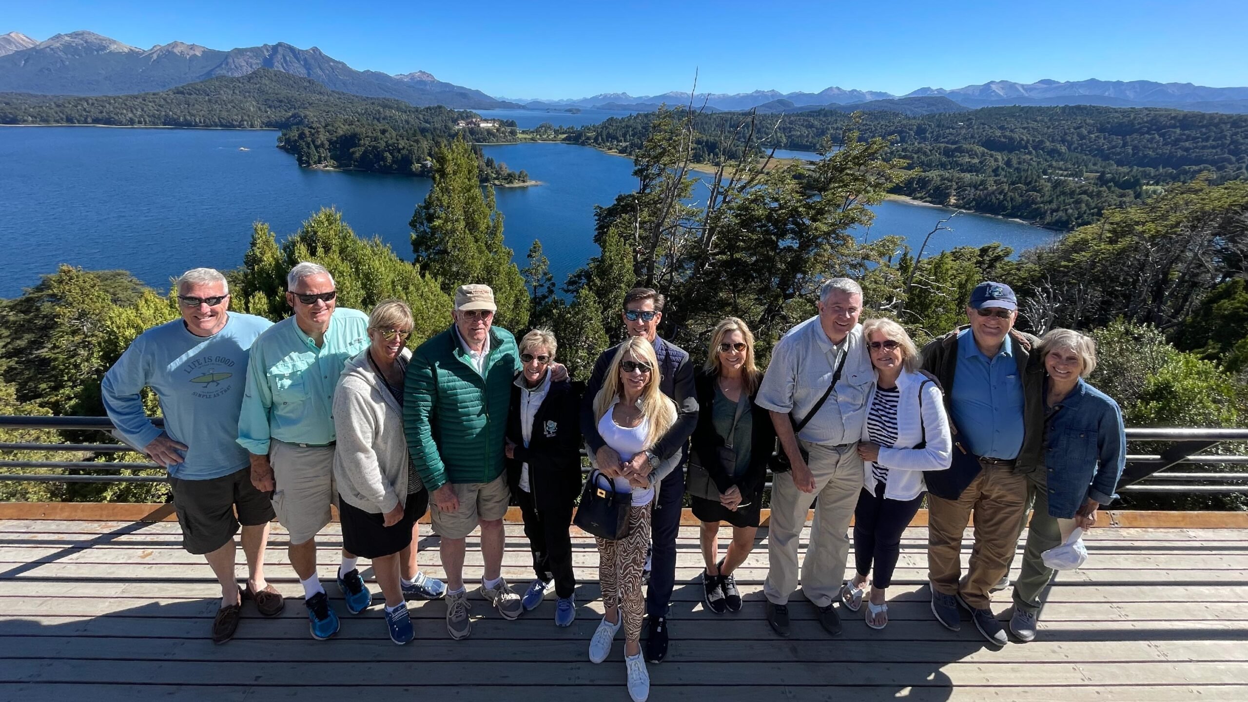 A group of travelers poses in front of Lake Nahuel Huapi in Bariloche, Argentina. They stand on a pebbled shore with the vast, serene lake and surrounding forested mountains in the background.