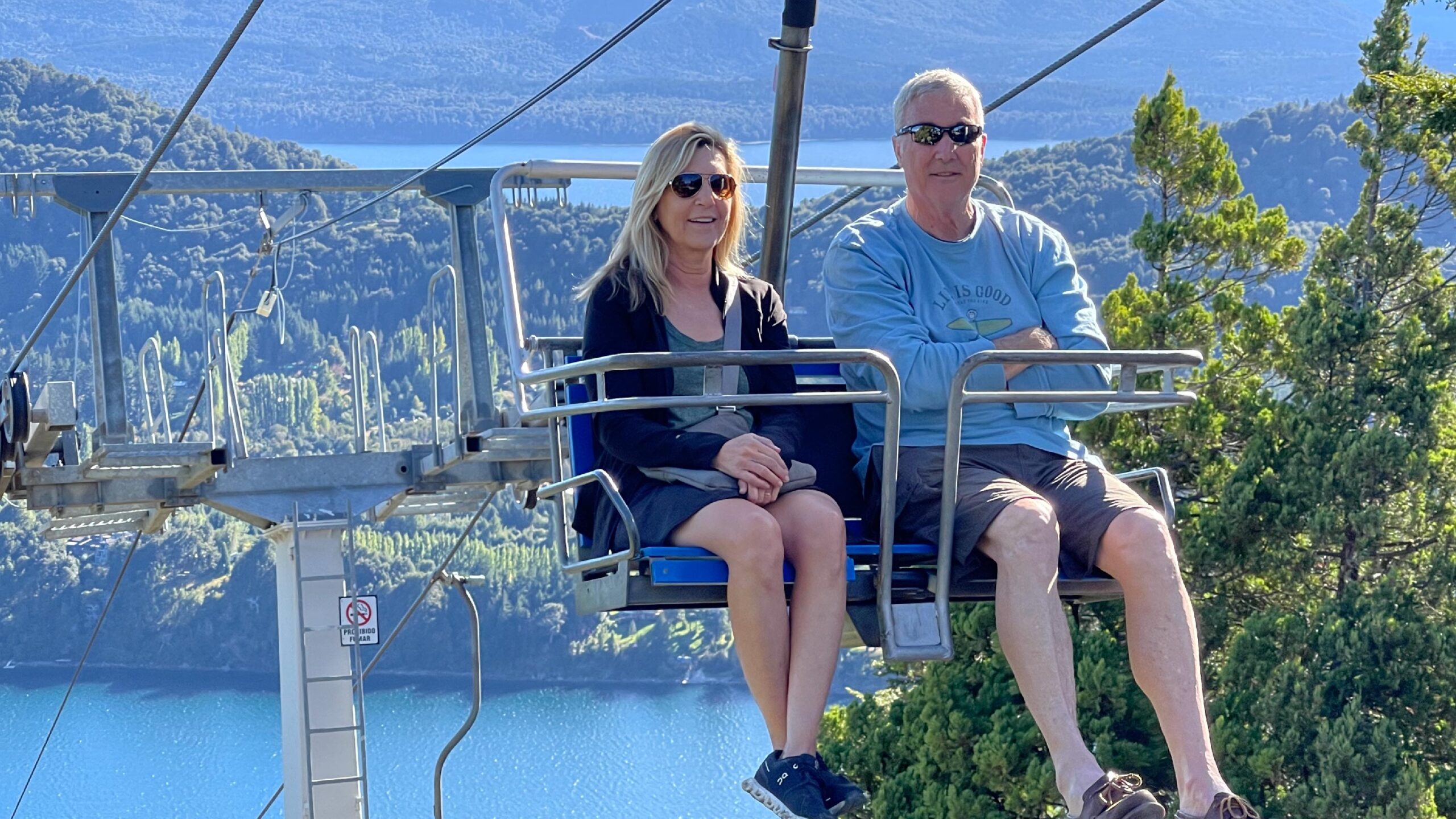 A couple of travelers poses inside a cable car in Bariloche, Argentina, with the panoramic view of the mountainous landscape and Lake Nahuel Huapi below.