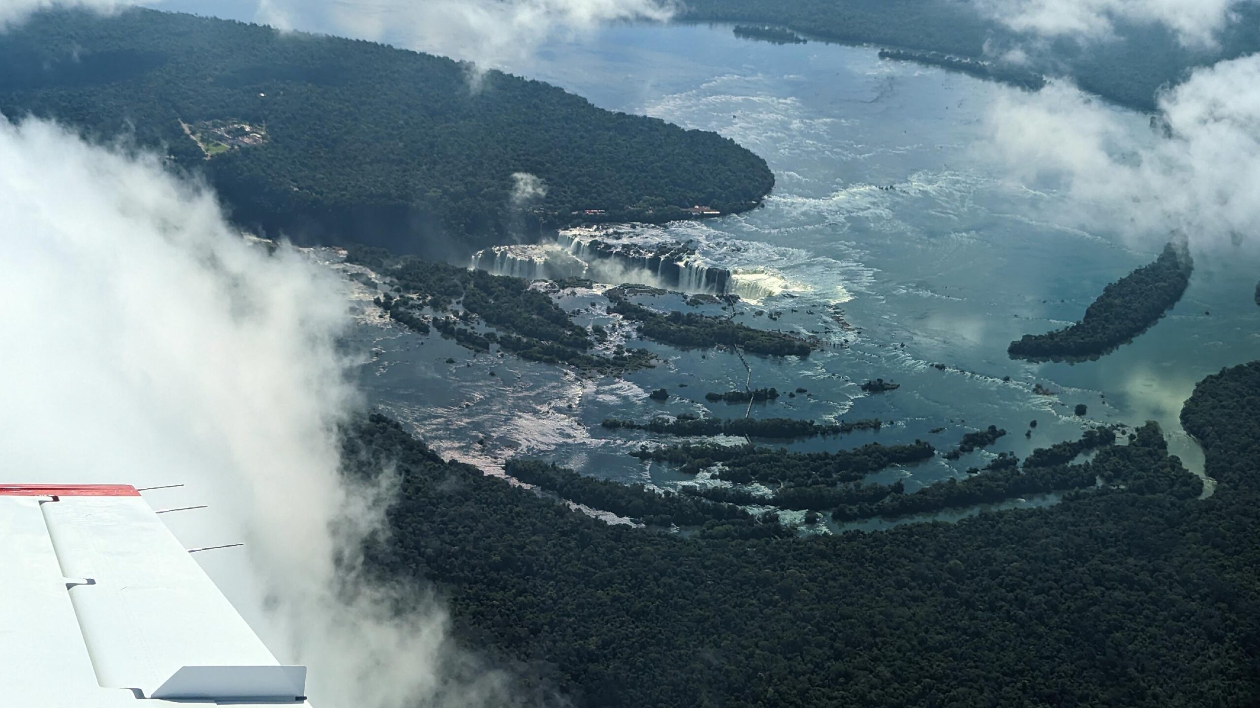 An aerial view of Iguazu Falls in Brazil reveals the stunning grandeur of the waterfalls. The image shows a series of powerful cascades surrounded by lush, dense rainforest.