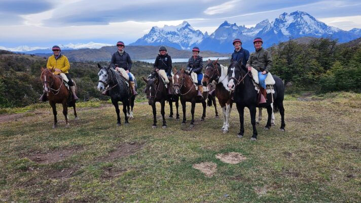 A group of travelers poses on horseback during a riding experience in Patagonia, Chile, with a backdrop of dramatic mountains, lush green fields, and a clear blue sky.