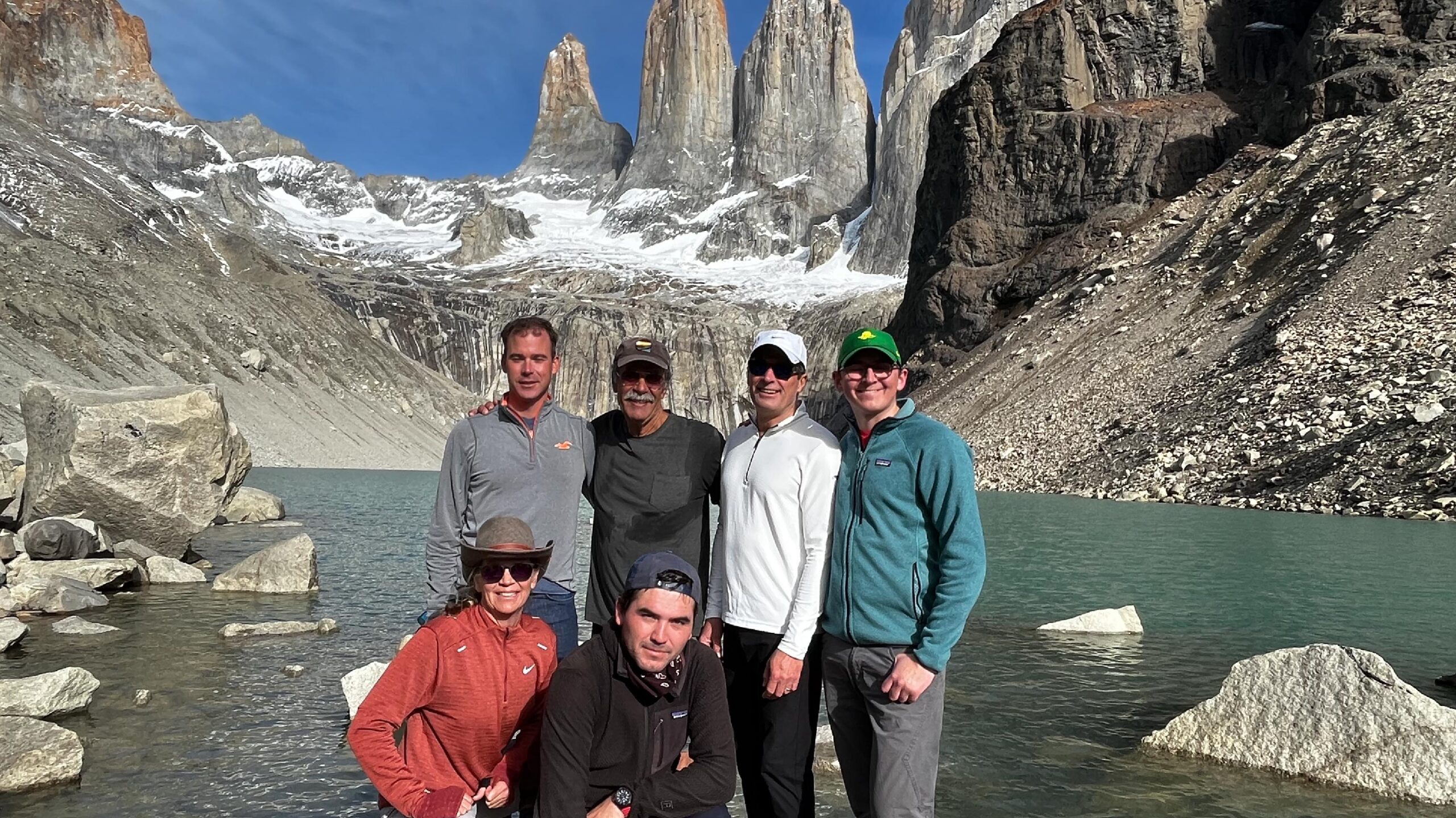 A group of travelers poses in front of the majestic Torres del Paine in Patagonia, Chile. They are bundled up in outdoor gear, smiling and standing on a rocky terrain with the towering granite peaks and snow-capped mountains of Torres del Paine National Park in the background under a clear blue sky.