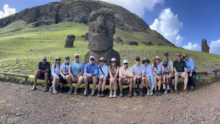 A group of travelers is sitting in front of a large Moai statue on Easter Island, Chile. The statue, carved from stone, stands tall against a backdrop of lush green grass and a partly cloudy sky.